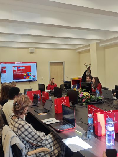 A meeting room with participants seated around a table, laptops open, and a presentation displayed on a screen. Red gift bags are visible.