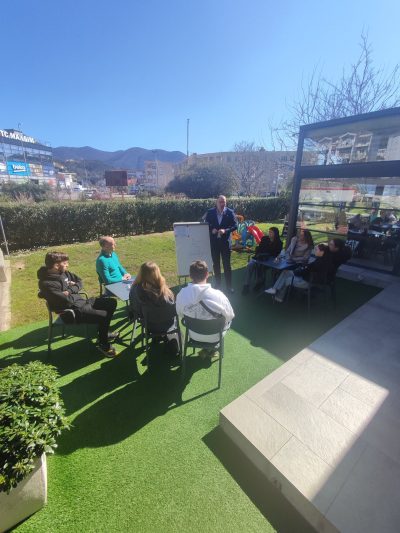 A group of people is seated outdoors, listening to a speaker in front of a whiteboard, with mountains in the background.